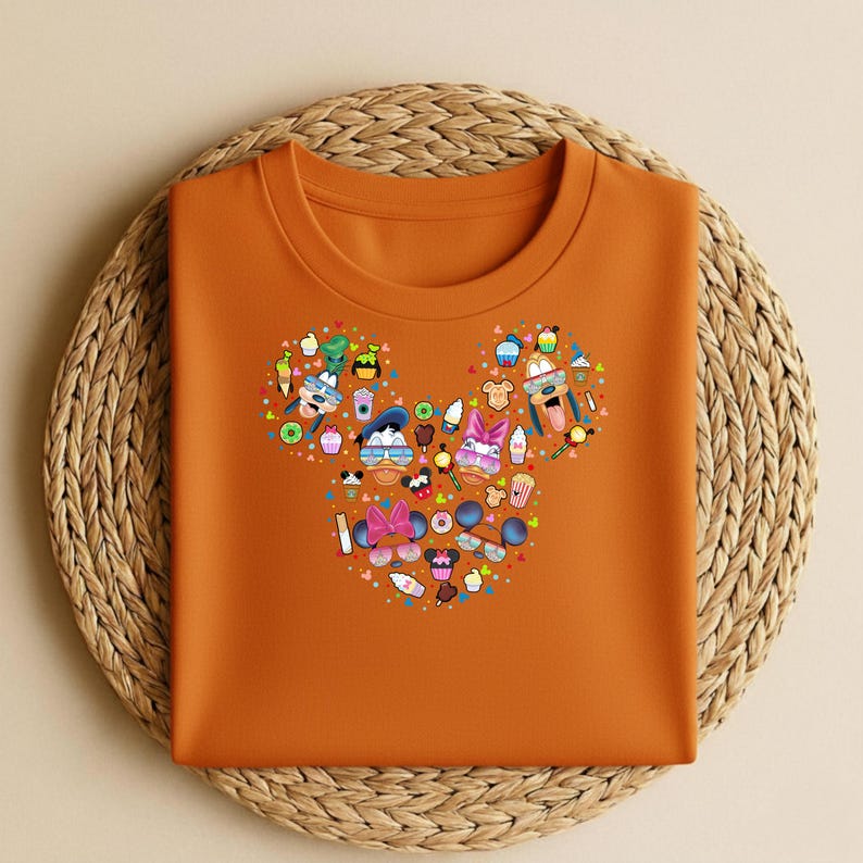 May include: An orange crew neck t-shirt with a Mickey Mouse head design composed of cartoon characters, food, and colourful shapes. The design includes ice cream cones, cupcakes, and sunglasses. The shirt is folded on a woven, circular placemat.