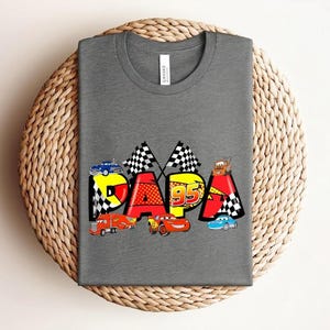 Puede incluir: Una camiseta gris con la palabra "PAPA" en letras coloridas de estilo caricatura. Las letras est&aacute;n decoradas con coches de carreras, banderas a cuadros y el n&uacute;mero "95". La camiseta est&aacute; colocada sobre una superficie circular tejida.