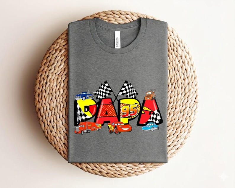 Puede incluir: Una camiseta gris con la palabra "PAPA" en letras coloridas de estilo caricatura. Las letras est&aacute;n decoradas con coches de carreras, banderas a cuadros y el n&uacute;mero "95". La camiseta est&aacute; colocada sobre una superficie circular tejida.