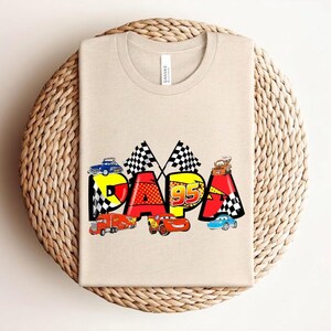 Puede incluir: Una camiseta beige con la palabra "PAPA" en letras coloridas de estilo caricatura, adornada con gr&aacute;ficos de coches de carreras. El dise&ntilde;o incluye banderas a cuadros y el n&uacute;mero "95". La camiseta se muestra sobre una superficie tejida.