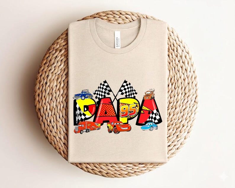Puede incluir: Una camiseta beige con la palabra "PAPA" en letras coloridas de estilo caricatura, adornada con gr&aacute;ficos de coches de carreras. El dise&ntilde;o incluye banderas a cuadros y el n&uacute;mero "95". La camiseta se muestra sobre una superficie tejida.