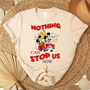 Puede incluir: Camiseta beige con Mickey y Minnie Mouse en un coche rojo y el texto "NOTHING CAN STOP US NOW" en rojo y negro. La camiseta tiene cuello redondo y mangas cortas.