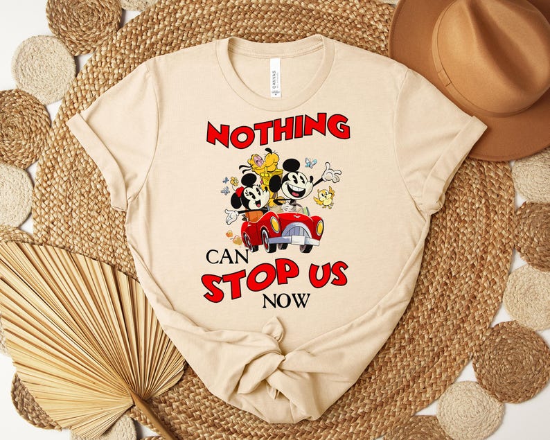 Puede incluir: Camiseta beige con Mickey y Minnie Mouse en un coche rojo y el texto "NOTHING CAN STOP US NOW" en rojo y negro. La camiseta tiene cuello redondo y mangas cortas.