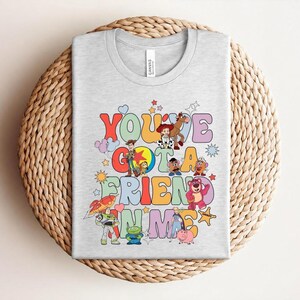 May include: A light grey t-shirt with the words "You've Got a Friend in Me" in colourful, retro lettering. The design features cartoon characters and stars, giving a playful and nostalgic feel.