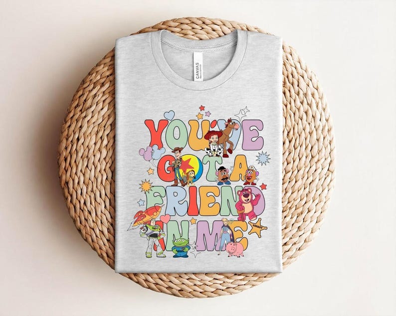 May include: A light grey t-shirt with the words "You've Got a Friend in Me" in colourful, retro lettering. The design features cartoon characters and stars, giving a playful and nostalgic feel.
