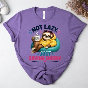 May include: A lilac t-shirt with a sloth graphic wearing sunglasses and a yellow hoodie, holding a coffee cup. The text reads "NOT LAZY. JUST SAVING ENERGY." and "NOT FAST, NOT FURIOUS, JUST ME."