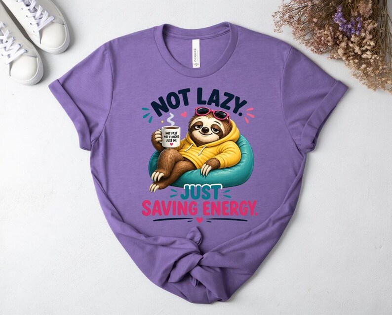 May include: A lilac t-shirt with a sloth graphic wearing sunglasses and a yellow hoodie, holding a coffee cup. The text reads "NOT LAZY. JUST SAVING ENERGY." and "NOT FAST, NOT FURIOUS, JUST ME."