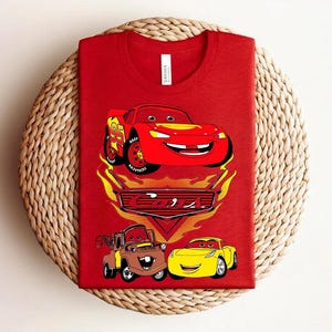 May include: Red t-shirt featuring a graphic of Lightning McQueen from the movie Cars, with the number 95 on the side. The design includes other characters and the Cars logo. The shirt is displayed on a woven surface.