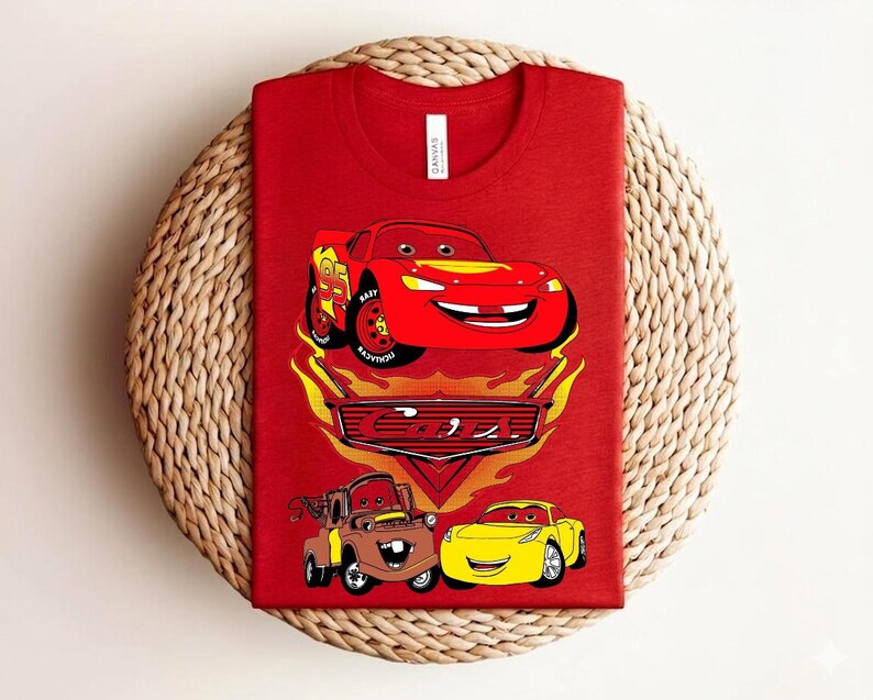May include: Red t-shirt featuring a graphic of Lightning McQueen from the movie Cars, with the number 95 on the side. The design includes other characters and the Cars logo. The shirt is displayed on a woven surface.