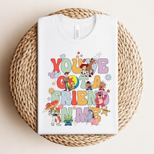 May include: White t-shirt with a colourful graphic design featuring the text "You've Got a Friend in Me" and cartoon characters. The design includes stars, clouds, and a rocket ship. The shirt is folded on a woven surface.