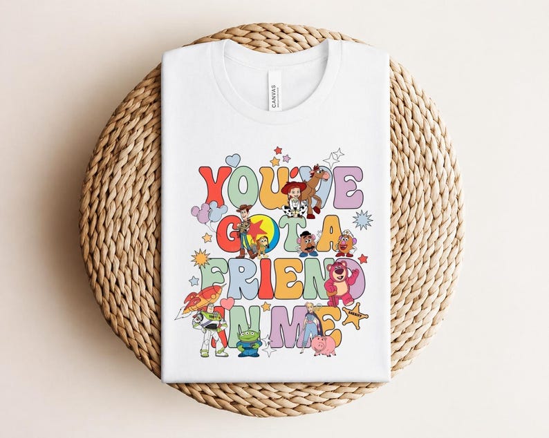 May include: White t-shirt with a colourful graphic design featuring the text "You've Got a Friend in Me" and cartoon characters. The design includes stars, clouds, and a rocket ship. The shirt is folded on a woven surface.