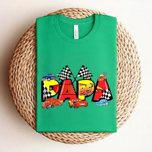 Puede incluir: Una camiseta verde con la palabra "PAPA" en un dise&ntilde;o colorido con coches de carreras y banderas a cuadros. El n&uacute;mero "95" tambi&eacute;n est&aacute; incorporado en el dise&ntilde;o. La camiseta est&aacute; hecha de un material suave.