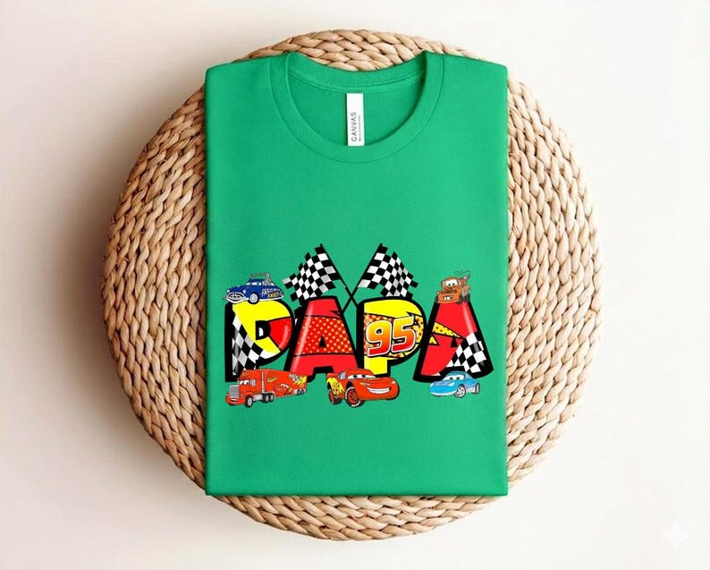 Puede incluir: Una camiseta verde con la palabra "PAPA" en un dise&ntilde;o colorido con coches de carreras y banderas a cuadros. El n&uacute;mero "95" tambi&eacute;n est&aacute; incorporado en el dise&ntilde;o. La camiseta est&aacute; hecha de un material suave.