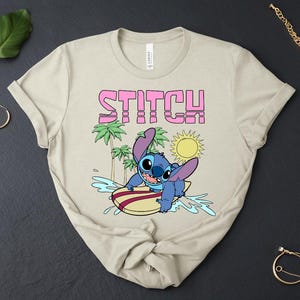 May include: Beige t-shirt featuring the word "STITCH" in pink block letters above a cartoon image of Stitch surfing. The graphic includes palm trees, a sun, and water splashes. The shirt is made of a soft material.