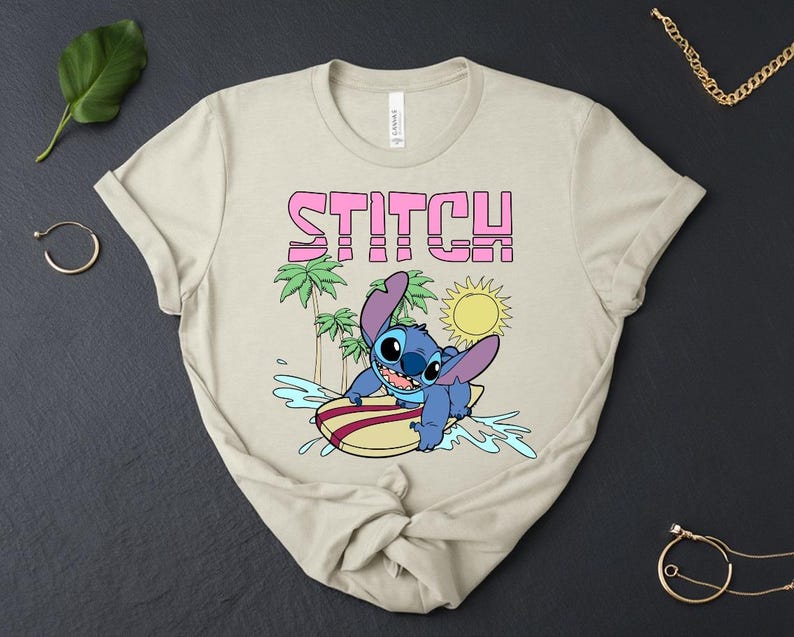 May include: Beige t-shirt featuring the word "STITCH" in pink block letters above a cartoon image of Stitch surfing. The graphic includes palm trees, a sun, and water splashes. The shirt is made of a soft material.