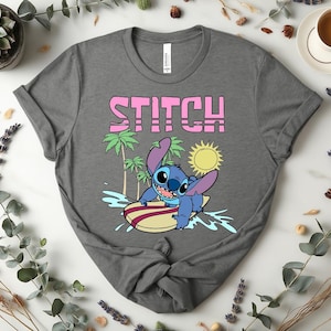 May include: A gray t-shirt featuring the word "STITCH" in pink block letters above a cartoon image of Stitch surfing on a surfboard. The design includes palm trees, a sun, and water splashes.