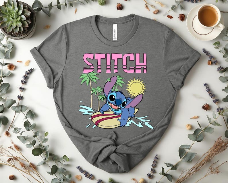 May include: A gray t-shirt featuring the word "STITCH" in pink block letters above a cartoon image of Stitch surfing on a surfboard. The design includes palm trees, a sun, and water splashes.