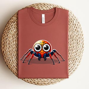 Pu&ograve; includere: Una maglietta color ruggine con un disegno di ragno dei cartoni animati. Il ragno ha una testa colorata e grandi occhi, con zampe rosse e blu. La maglietta &egrave; piegata su una superficie circolare intrecciata.
