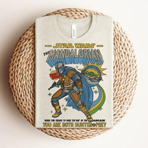K&ouml;nnte beinhalten: Beiges T-Shirt mit einem farbenfrohen Grafikdesign von Star Wars The Mandalorian. Das Design enth&auml;lt den Text "STAR WARS THE MANDALORIAN" und das Zitat "YOU ARE BOTH HUNTER PREY". Die Illustration zeigt den Mandalorianer und Grogu.