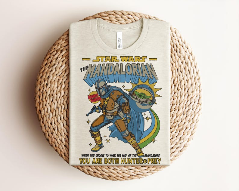 K&ouml;nnte beinhalten: Beiges T-Shirt mit einem farbenfrohen Grafikdesign von Star Wars The Mandalorian. Das Design enth&auml;lt den Text "STAR WARS THE MANDALORIAN" und das Zitat "YOU ARE BOTH HUNTER PREY". Die Illustration zeigt den Mandalorianer und Grogu.
