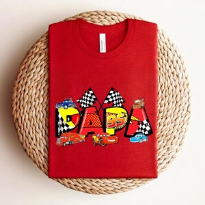 Puede incluir: Camiseta roja con la palabra "PAPA" en un dise&ntilde;o colorido con coches de carreras y banderas a cuadros. El n&uacute;mero "95" tambi&eacute;n est&aacute; incluido en el dise&ntilde;o. La camiseta est&aacute; hecha de un material suave.