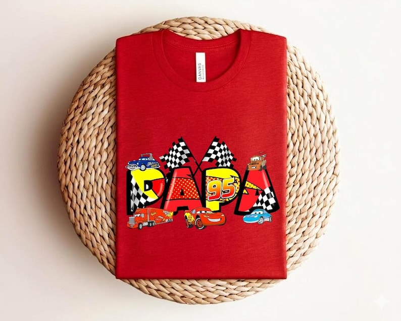 Puede incluir: Camiseta roja con la palabra "PAPA" en un dise&ntilde;o colorido con coches de carreras y banderas a cuadros. El n&uacute;mero "95" tambi&eacute;n est&aacute; incluido en el dise&ntilde;o. La camiseta est&aacute; hecha de un material suave.