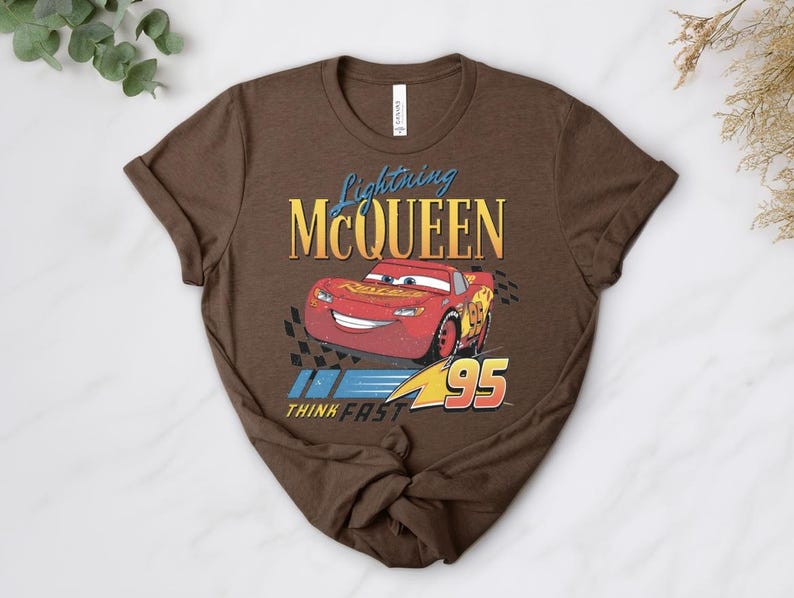 May include: A brown t-shirt with a graphic of Lightning McQueen, a red race car, and the text "Lightning McQueen" above the car and "Think Fast" below. The number "95" is displayed.