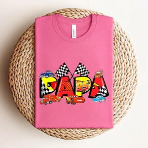 Puede incluir: Una camiseta rosa con la palabra "DAPA" en letras coloridas, con coches de carreras y banderas a cuadros. El n&uacute;mero "95" se muestra de forma destacada. La camiseta est&aacute; colocada sobre una superficie circular tejida.