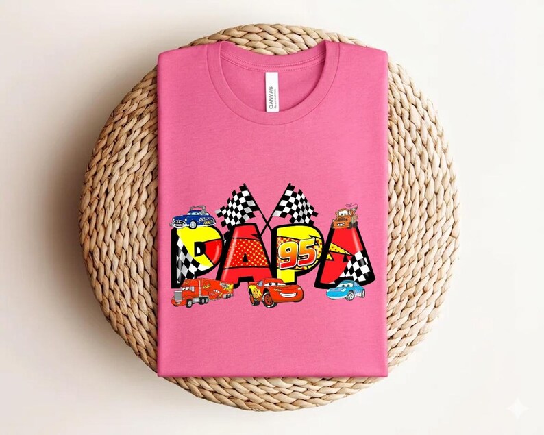 Puede incluir: Una camiseta rosa con la palabra "DAPA" en letras coloridas, con coches de carreras y banderas a cuadros. El n&uacute;mero "95" se muestra de forma destacada. La camiseta est&aacute; colocada sobre una superficie circular tejida.