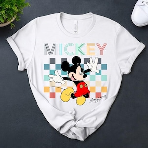 May include: White t-shirt featuring a colorful graphic of Mickey Mouse. The design includes the word "MICKEY" in various colors above the character. Mickey is depicted with his arms outstretched, wearing red shorts and yellow shoes.