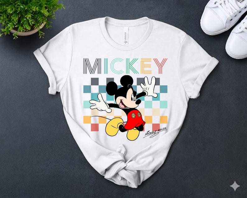May include: White t-shirt featuring a colorful graphic of Mickey Mouse. The design includes the word "MICKEY" in various colors above the character. Mickey is depicted with his arms outstretched, wearing red shorts and yellow shoes.
