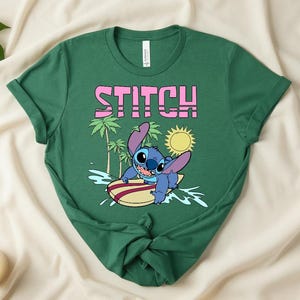 May include: Green t-shirt featuring the word "STITCH" in pink block letters above a cartoon image of Stitch surfing on a yellow and red surfboard. The design includes palm trees, a yellow sun, and blue water.