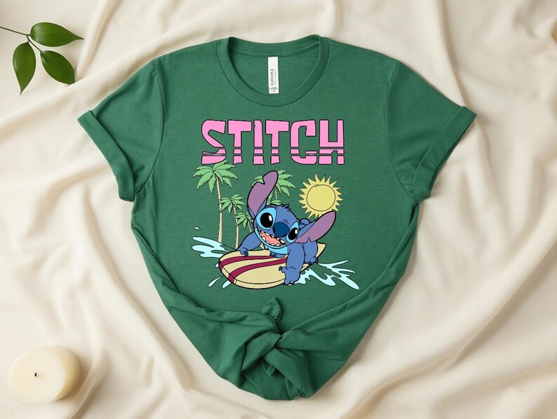 May include: Green t-shirt featuring the word "STITCH" in pink block letters above a cartoon image of Stitch surfing on a yellow and red surfboard. The design includes palm trees, a yellow sun, and blue water.