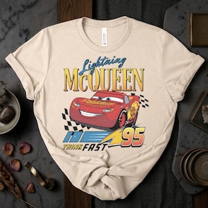 May include: Beige t-shirt featuring Lightning McQueen from the movie Cars. The design includes the text "Lightning McQueen" above a red race car with the number 95, and the words "Think Fast".