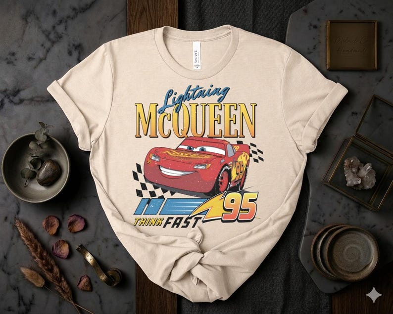 May include: Beige t-shirt featuring Lightning McQueen from the movie Cars. The design includes the text "Lightning McQueen" above a red race car with the number 95, and the words "Think Fast".