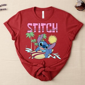 May include: Red t-shirt featuring the word "STITCH" in pink block letters. The shirt has a graphic of the character Stitch surfing on a surfboard, with palm trees and a sun in the background. Sunglasses are on the top right.