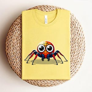 Pu&ograve; includere: T-shirt giallo senape con un'illustrazione di ragno dei cartoni animati. Il ragno ha un corpo rosso, blu e giallo, grandi occhi e zampe rosse e nere. La maglietta &egrave; piegata su un tappetino circolare intrecciato.