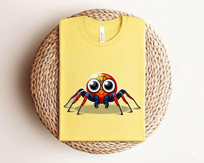 Pu&ograve; includere: T-shirt giallo senape con un'illustrazione di ragno dei cartoni animati. Il ragno ha un corpo rosso, blu e giallo, grandi occhi e zampe rosse e nere. La maglietta &egrave; piegata su un tappetino circolare intrecciato.