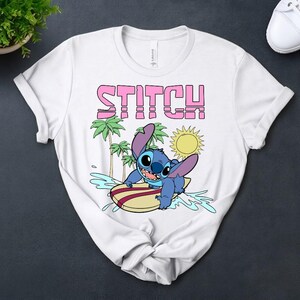 May include: White t-shirt featuring the word "STITCH" in pink block letters above a cartoon image of Stitch surfing. The blue character is on a surfboard with palm trees and a sun in the background. The shirt is made of a soft material.