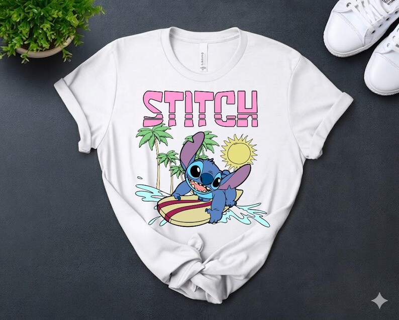 May include: White t-shirt featuring the word "STITCH" in pink block letters above a cartoon image of Stitch surfing. The blue character is on a surfboard with palm trees and a sun in the background. The shirt is made of a soft material.
