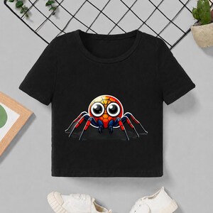 Pu&ograve; includere: Top corto nero con una grafica di ragno dei cartoni animati colorata. Il ragno ha un corpo rotondo con dettagli rossi, gialli e arancioni, grandi occhi e zampe rosse e nere. La maglietta &egrave; abbinata a scarpe di tela bianche.