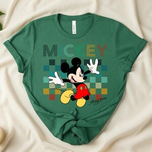 May include: Green t-shirt featuring Mickey Mouse with the word "MICKEY" in a colorful, pixelated design. Mickey is depicted with white gloves, a red shorts, and yellow shoes. The shirt has rolled-up sleeves.