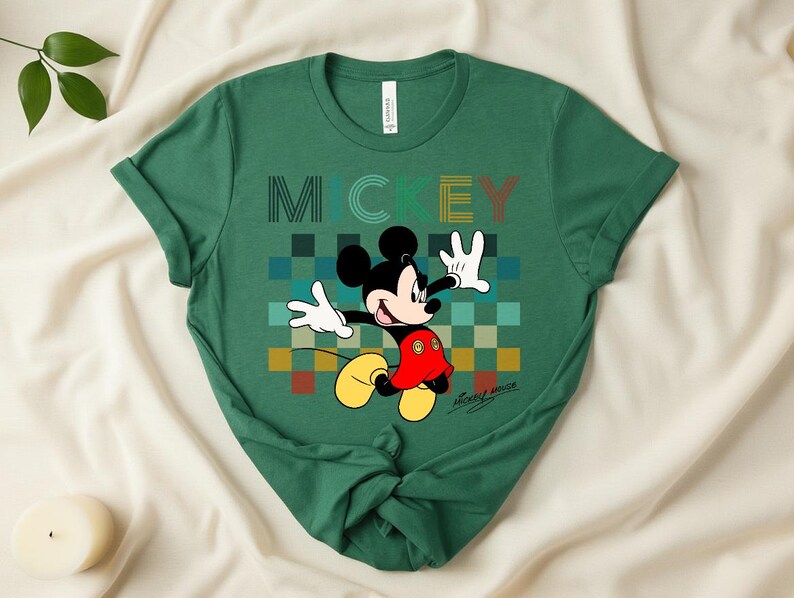 May include: Green t-shirt featuring Mickey Mouse with the word "MICKEY" in a colorful, pixelated design. Mickey is depicted with white gloves, a red shorts, and yellow shoes. The shirt has rolled-up sleeves.