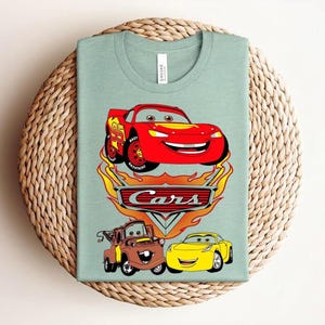 May include: A light green t-shirt featuring a graphic of the Cars movie characters. The design includes a red race car, a brown tow truck, and a yellow car, with the word "Cars" in a flame-themed logo.