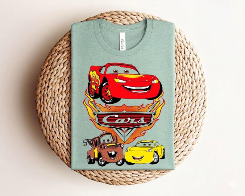 May include: A light green t-shirt featuring a graphic of the Cars movie characters. The design includes a red race car, a brown tow truck, and a yellow car, with the word "Cars" in a flame-themed logo.