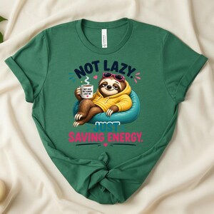 May include: A green t-shirt with the slogan "NOT LAZY. JUST SAVING ENERGY." features a cartoon sloth wearing sunglasses and a yellow hoodie, holding a coffee mug. The sloth is sitting on a blue beanbag chair.