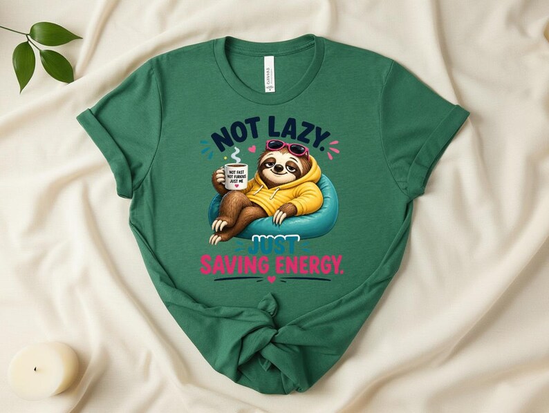 May include: A green t-shirt with the slogan "NOT LAZY. JUST SAVING ENERGY." features a cartoon sloth wearing sunglasses and a yellow hoodie, holding a coffee mug. The sloth is sitting on a blue beanbag chair.