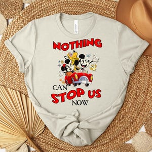 Puede incluir: Camiseta beige claro con un gr&aacute;fico colorido de Mickey y Minnie Mouse en un coche rojo, con el texto rojo "NOTHING CAN STOP US NOW". El dise&ntilde;o incluye mariposas y un p&aacute;jaro amarillo.