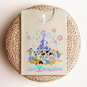 Może przedstawiać: Beżowa koszulka z kolorową grafiką postaci Disneya, w tym Myszki Miki, Myszki Minnie, Kaczora Donalda i Goofy'ego, na tle zamku z fajerwerkami. Tekst "Walt Disney World" jest wyświetlany pod postaciami.