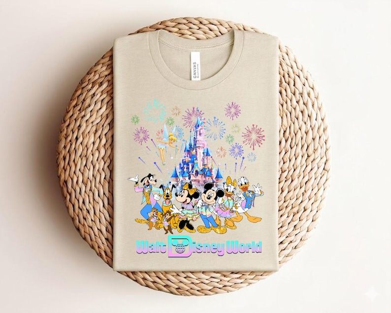 Może przedstawiać: Beżowa koszulka z kolorową grafiką postaci Disneya, w tym Myszki Miki, Myszki Minnie, Kaczora Donalda i Goofy'ego, na tle zamku z fajerwerkami. Tekst "Walt Disney World" jest wyświetlany pod postaciami.